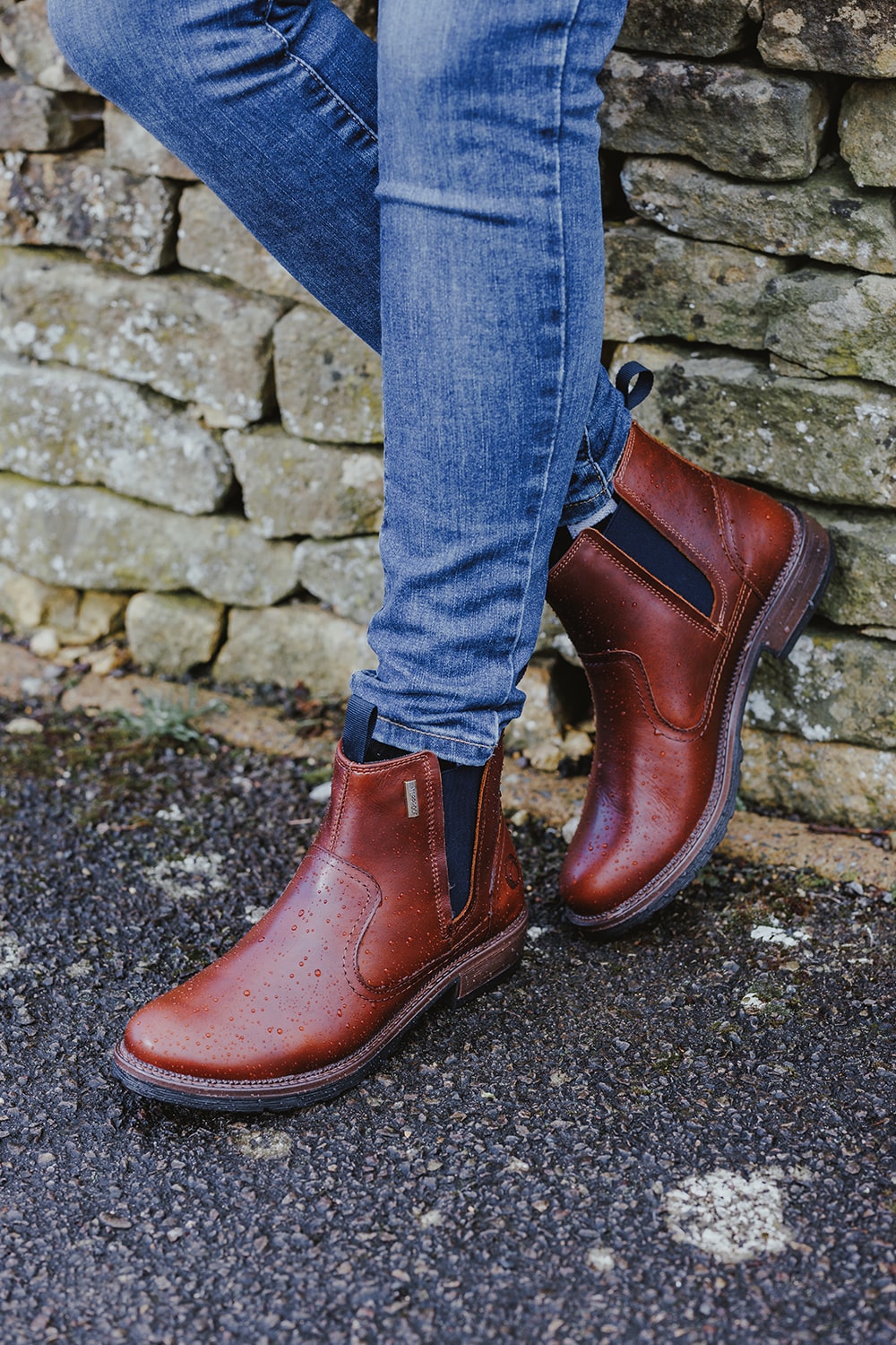 Close up image of a person stood in front of a dry stone wall wearing a pair of Cotswold Laverton boots