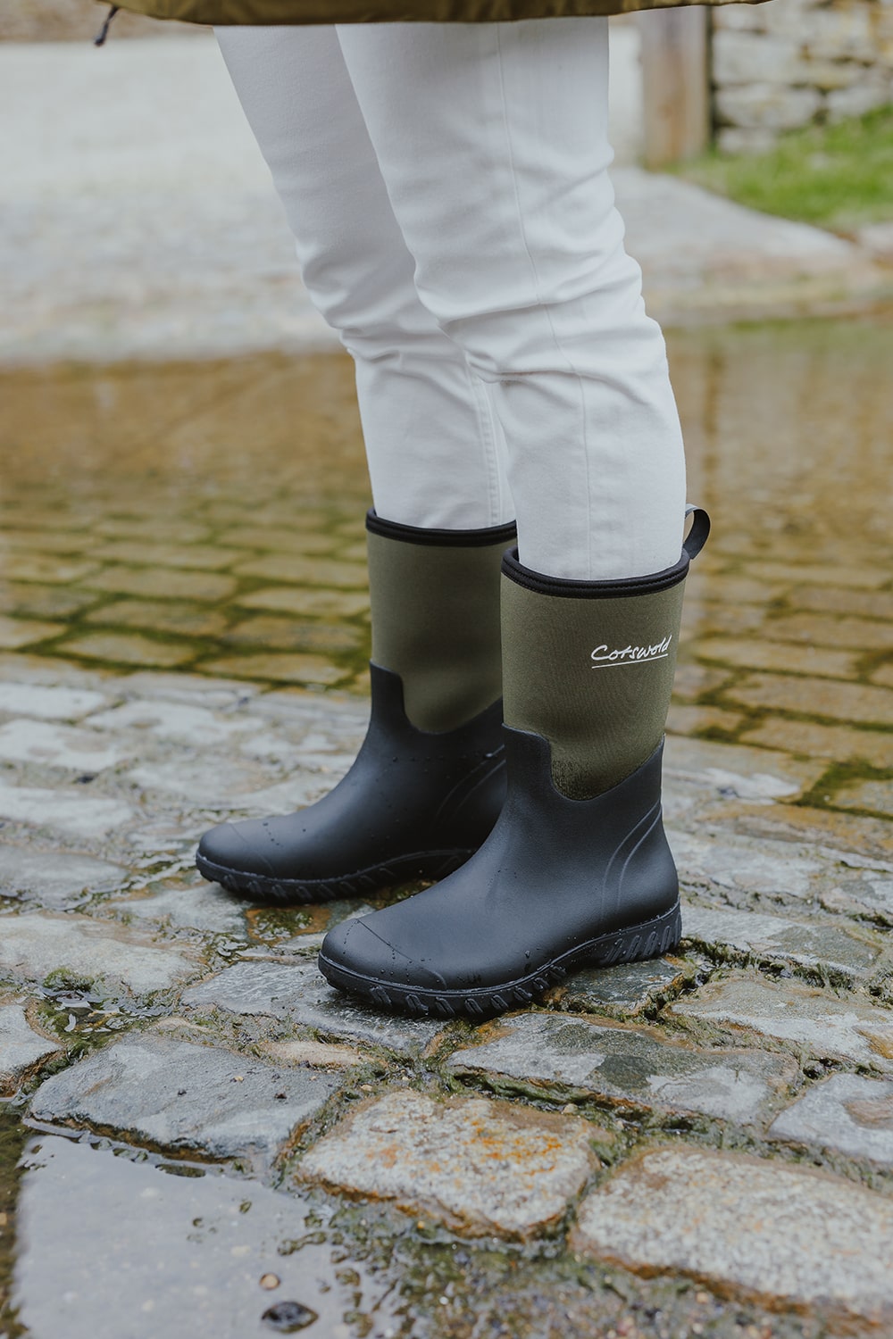 Close up image of a person stood on wet cobble stones, wearing a pair of Cotswold wellingtons