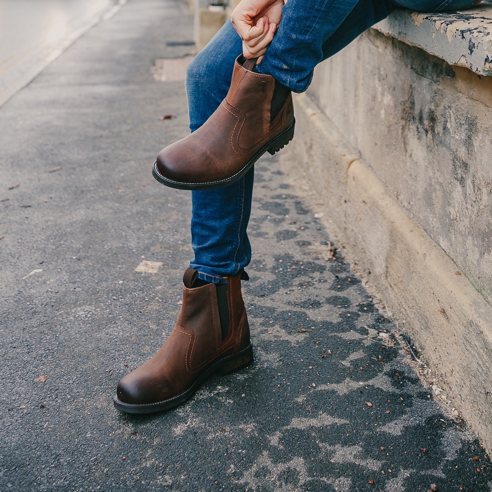 A man sat on a wall outside, putting on a pair of brown Cotswold Laverton Ankle Boots