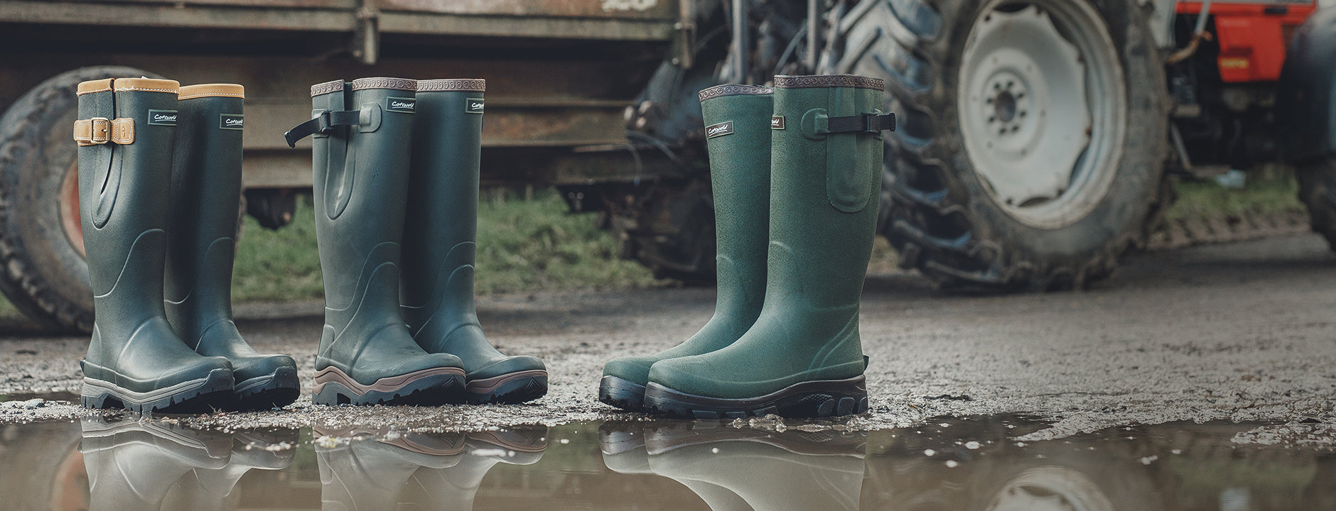 Three pairs of Cotswold wellingtons with reflections in the water and a tractor in the background